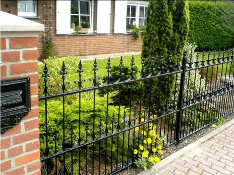 Clôture en fer forgé noir devant une maison, jardin avec arbustes et fleurs jaunes, pilier en briques, boîte aux lettres.